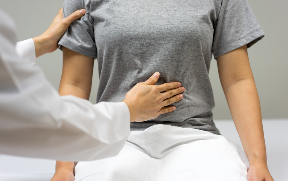 Female doctor are examining by abdominal palpation of female patient sitting in the bed within the clinic.