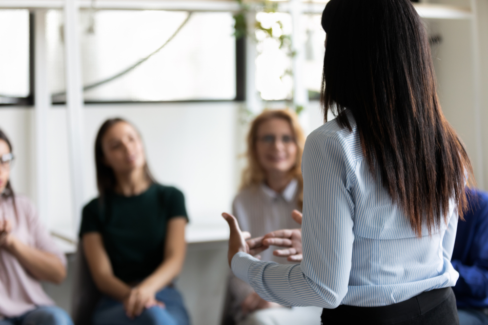 Rear back view female african ethnicity participant at group rehab session telling story share personal problem with associates or corporate staff gathered together listening business coach at seminar