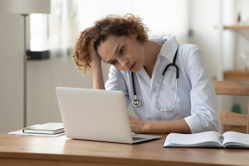 Stressed young female doctor looking at laptop reading bad news online worried of mistake at workplace. Unhappy professional physician in tension feeling tired solving computer problem at work.