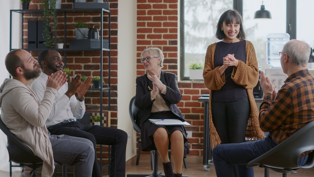 Happy adult celebrating recovery success with group of people clapping hands at aa meeting. Woman sharing achievement and progress at therapy, receiving applause from addiction patients.
