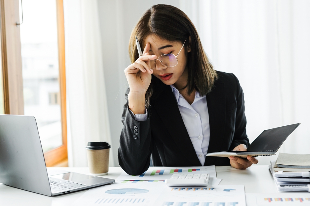 Attractive Asian short-haired businesswoman wearing glasses feels stressed as she works on financial documents at a desk in a modern office.
