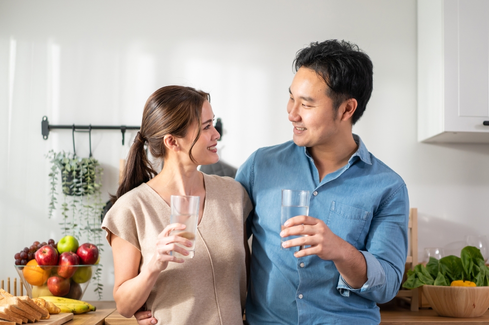 Asian attractive couple drinking a glass of water in kitchen at home. Young thirsty man and woman holding clean mineral natural in cup after waking up and sip in morning for health care in house.
