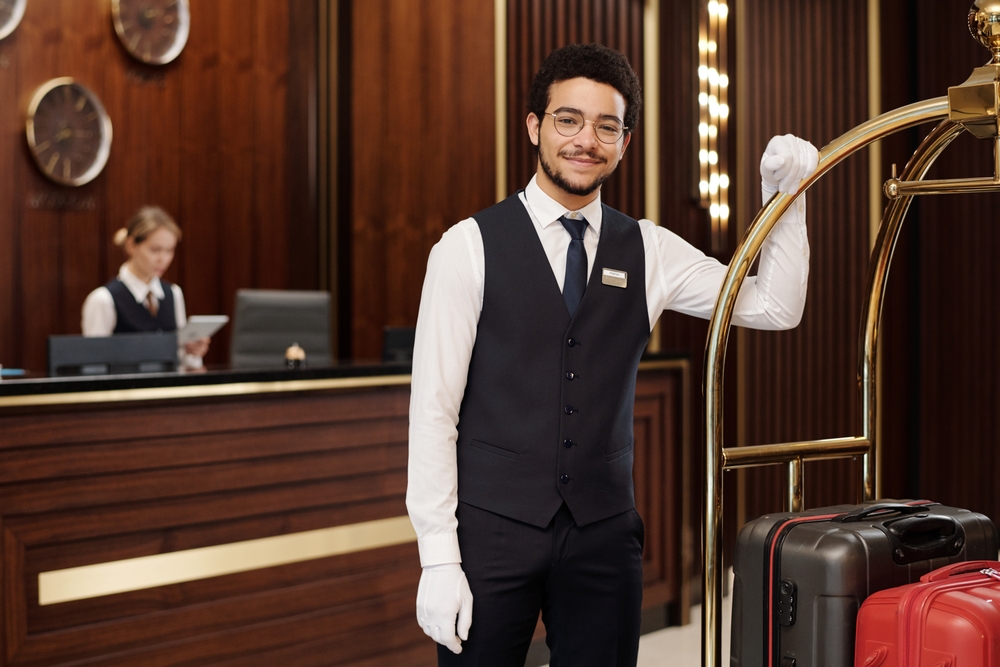 Young successful bellboy in elegant uniform and white gloves looking at camera while standing by cart with baggage of travelers