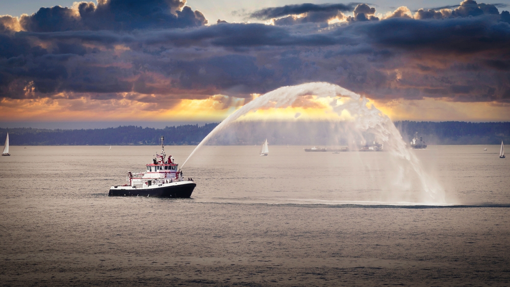 A tug boat is seen in the waters of a harbor, with water droplets being sprayed off its stern
