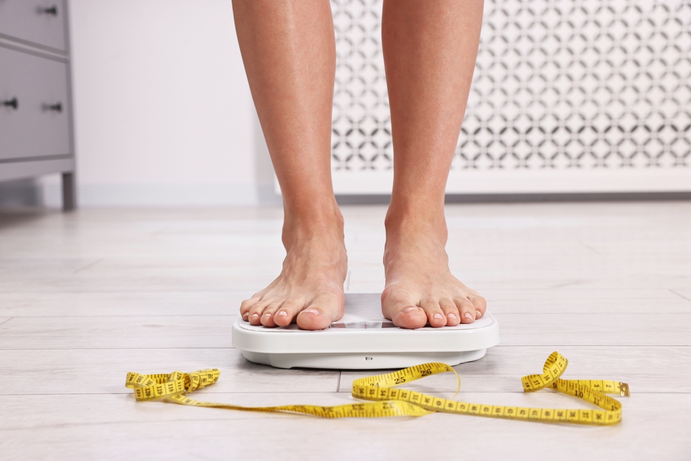 Woman standing on floor scale and measuring tape at home, closeup. Weight control
