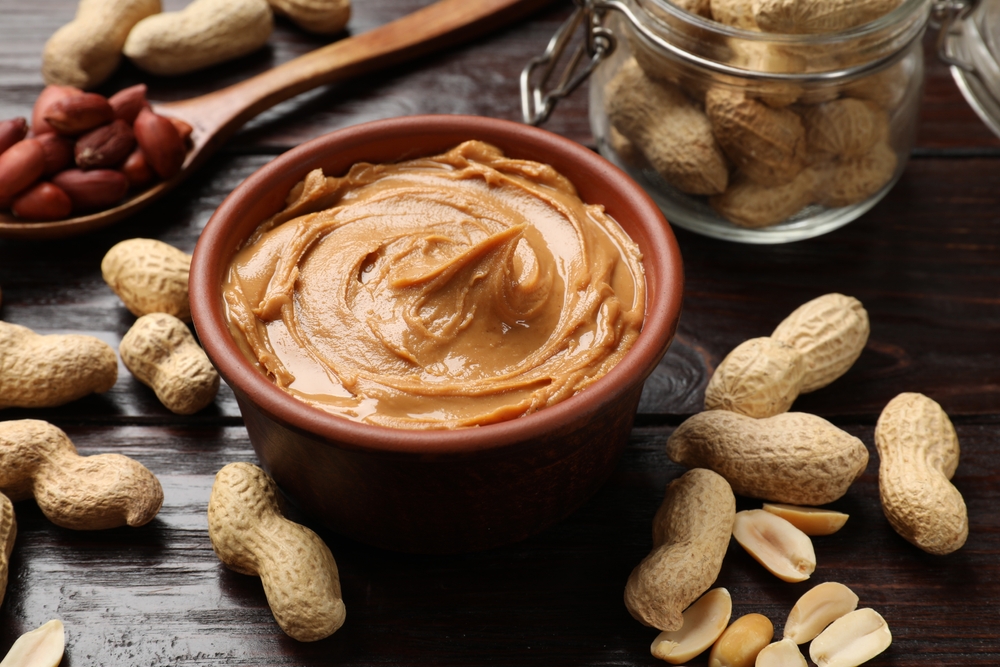 Tasty peanut butter in bowl, groundnuts, jar and spoon on wooden table, closeup
