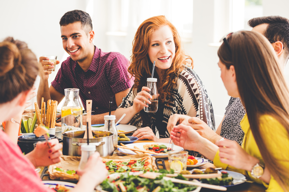 Group of mixed-race people celebrating vegan party at home, sitting at colorful table full of healthy snacks, salads and organic dishes
