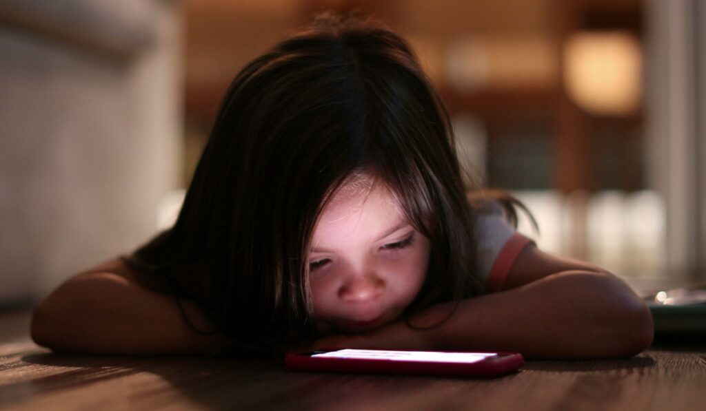 Small girl staring at cellphone screen at night. Cute kid lying on floor watching cellphone screen
