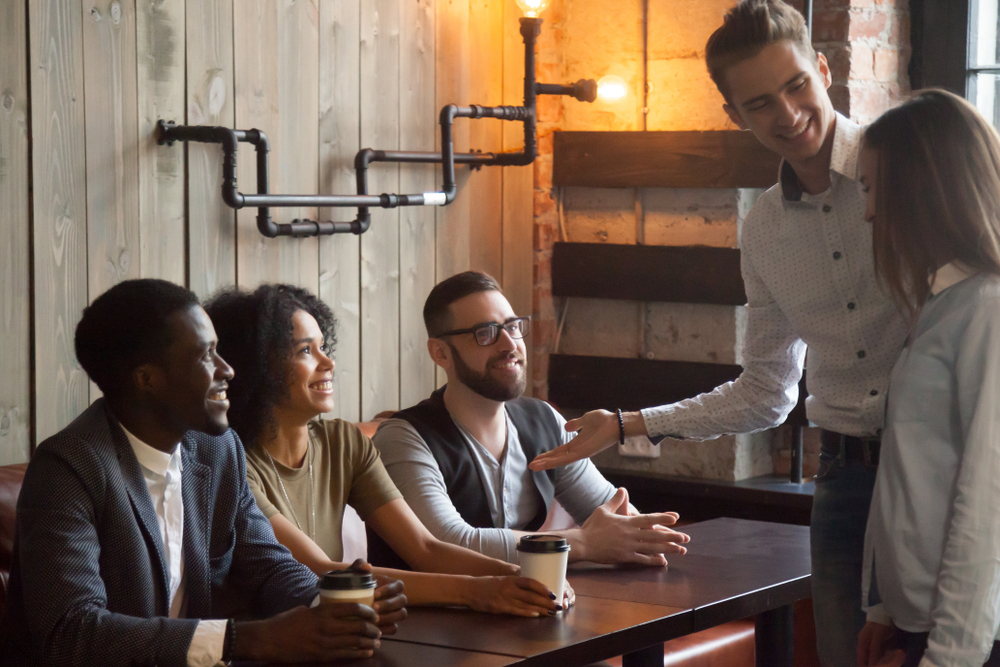 Smiling Caucasian boyfriend introducing multiracial friends to girlfriend at meeting in coffeeshop, millennial male acquainting diverse colleagues with female worker in cafe. First impression concept

