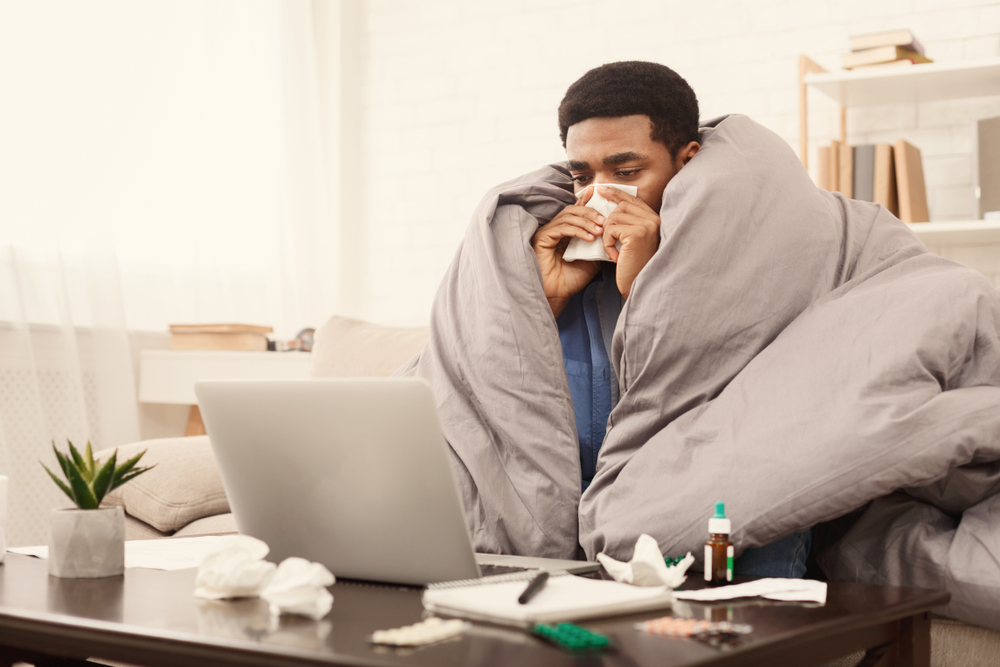 Sick black student working from home on laptop with homework assignment or university project, wrapped under blanket on couch, free space