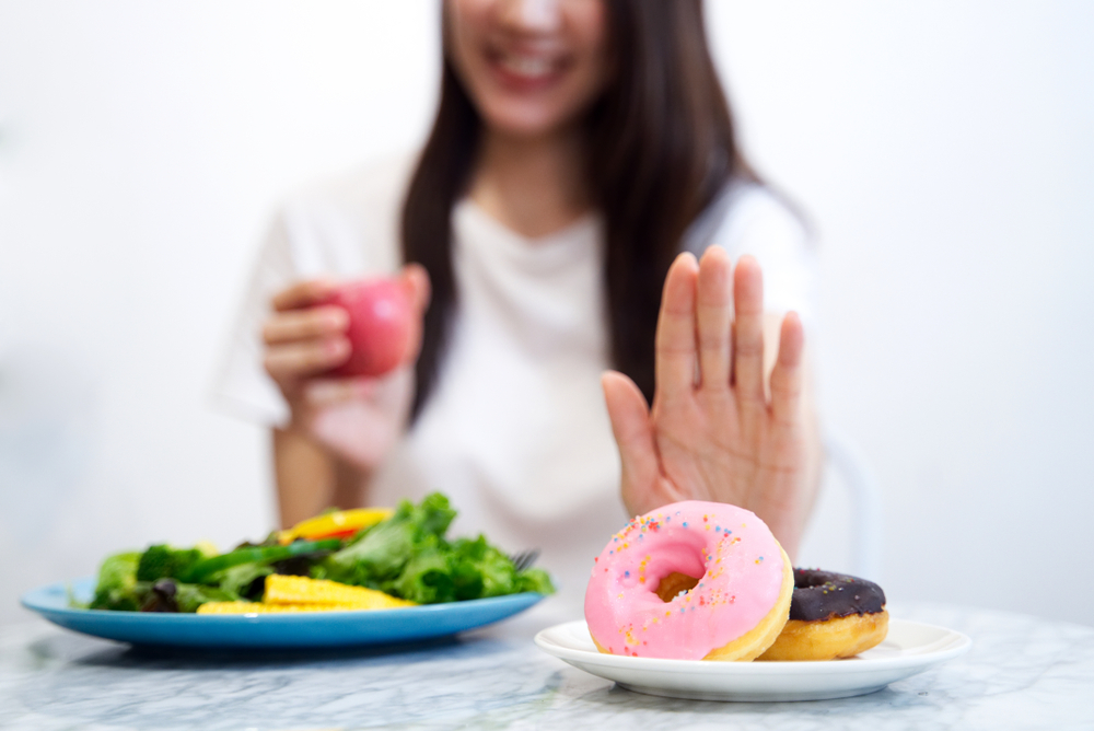 Young girl on dieting for good health concept. Close up female using hand reject junk food by pushing out her favorite sweet donuts and choose red apple and organic salad for good health at home.