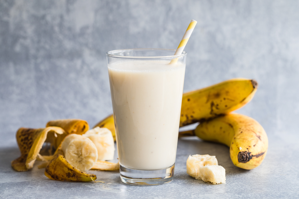 Banana smoothie of organic bananas. There is a high glass filled with banana smoothie in the centre, and bananas and banana peels around the drink. With an environmentally friendly paper straw.
