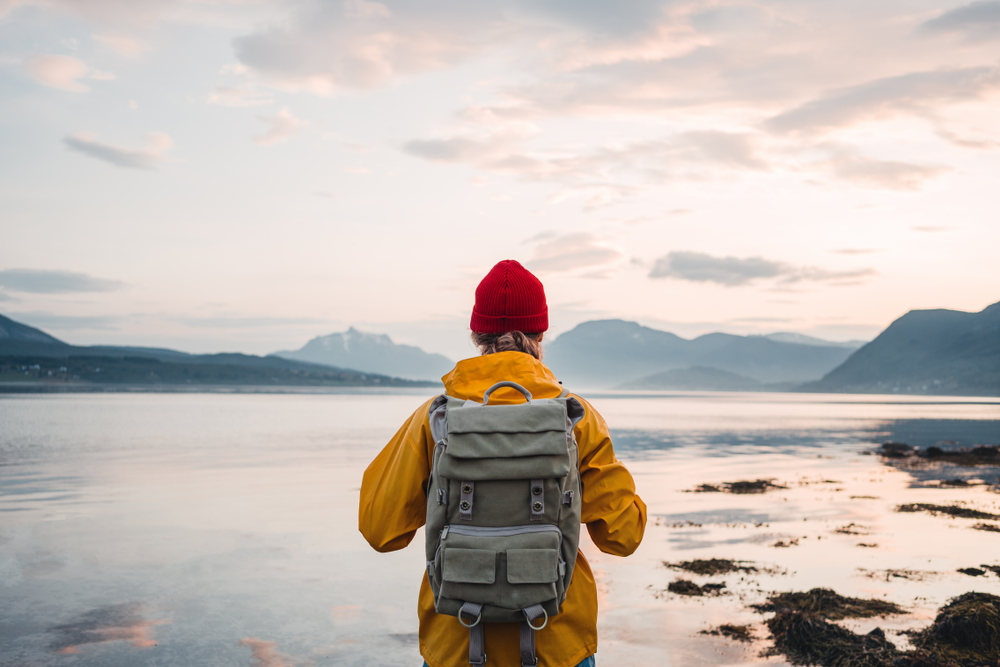 Back view of male tourist with rucksack standing on coast in front of great mountain massif while journey. Man traveler wearing yellow jacket with backpack explore Scandinavia nature. Wanderlust
