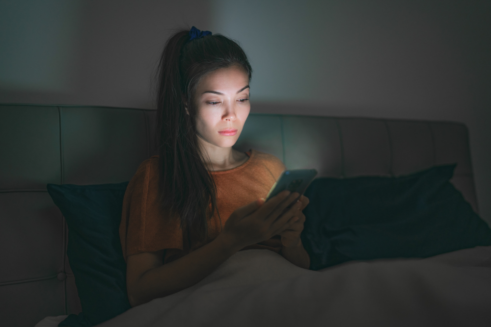 Phone in bed - tired asian woman staying up late at night using mobile cellphone addicted to social media smartphone unable to sleep awake from insomnia. Cell light lighting up face in the dark.