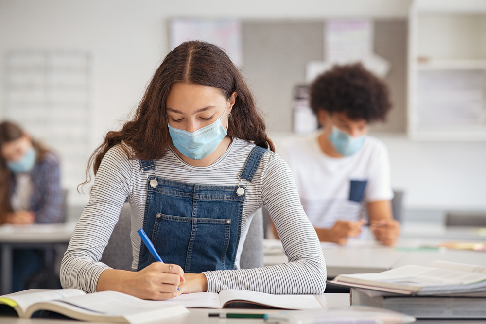 High school student taking notes while wearing face mask due to coronavirus emergency. Young woman sitting in class with their classmates and wearing surgical mask due to Covid-19 pandemic.