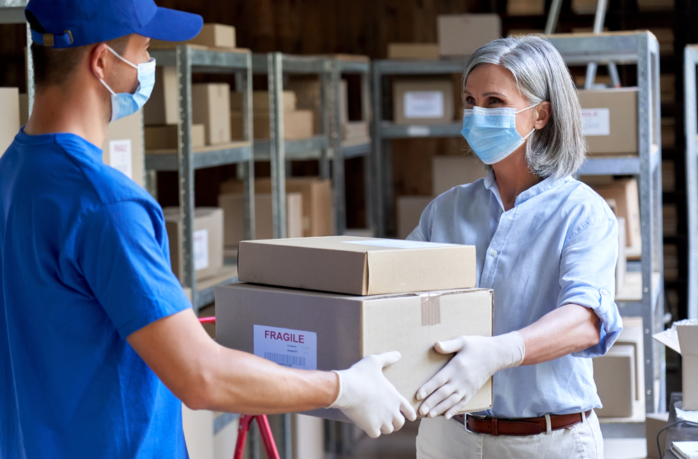 Female manager supervisor wearing face mask preparing fast drop shipping safe delivery giving parcels packages boxes to male courier taking ecommerce orders to deliver standing in warehouse storage.