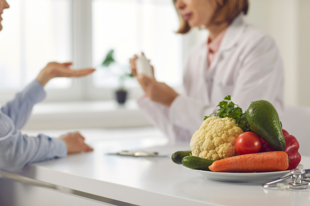 Doctor talking to patient and teaching her to take medicine, eat healthy food and stick to diet. Nutrition education and disease prevention concept. Blurred background, bowl of vegetables in close-up
