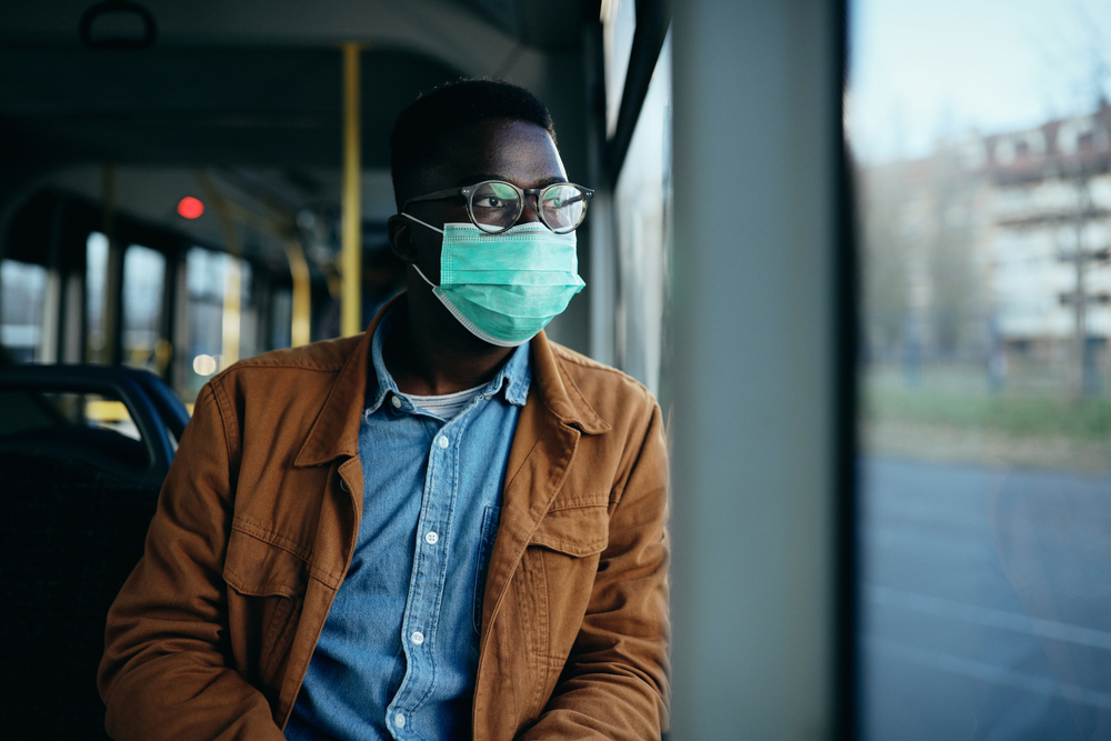 Pensive black man looking through the window and wearing face mask while commuting by bus during coronavirus pandemic.