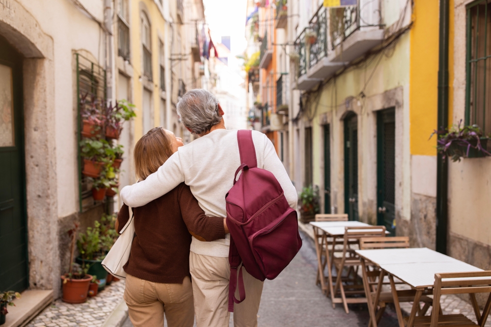 Vacation Tour Offer. Back View Of Senior Couple Hugging And Carrying Backpack Walking On Lisbon Street, Enjoying Sightseeing Outdoors. Travel And Tourism In Europe Concept
