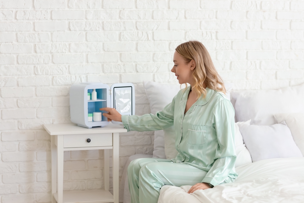 Beautiful young happy woman with small refrigerator for cosmetic products sitting in bedroom