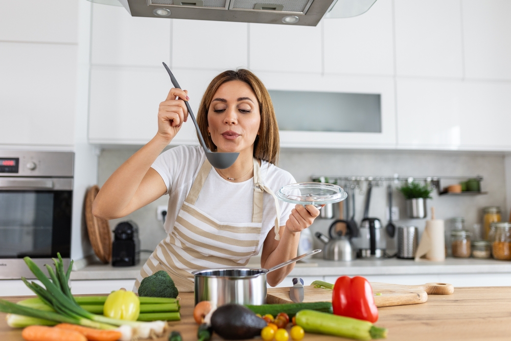Happy Young Woman Cooking Tasting Dinner In A Pot Standing In Modern Kitchen At Home. Housewife Preparing Healthy Food Smiling . Household And Nutrition. Dieting Recipes Concept
