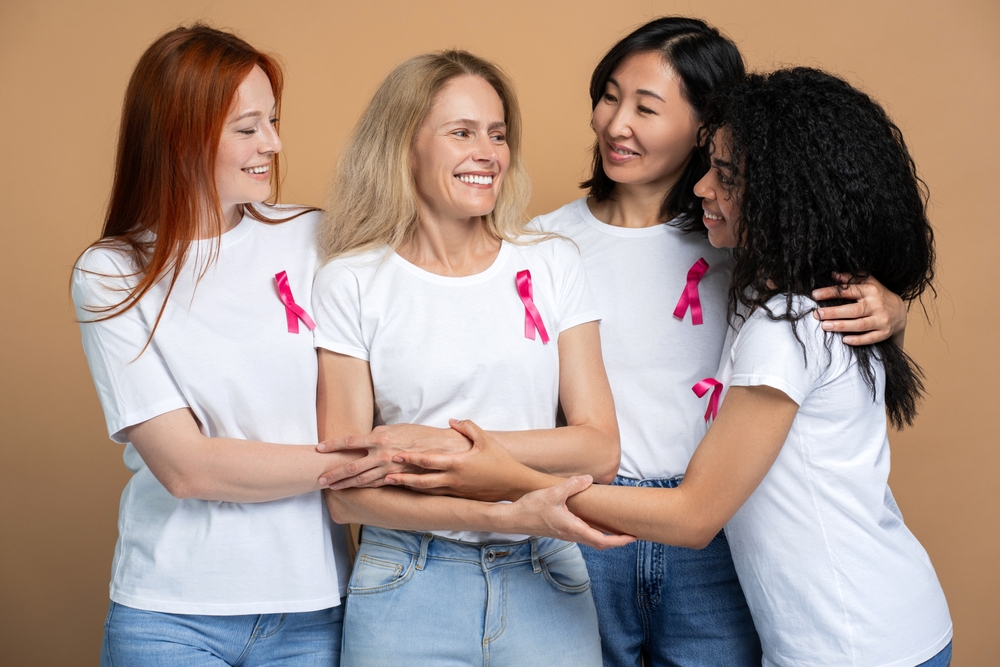 Four diverse women are smiling and showing their support for a friend battling breast cancer standing isolated on beige background. Support, health care concept