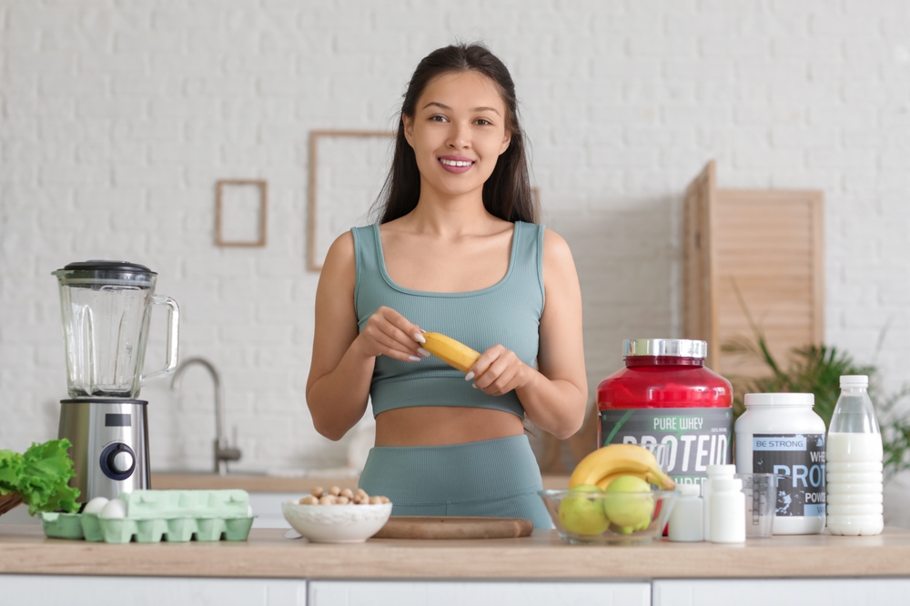Sporty young happy Asian woman with banana and ingredients for protein shake at home
