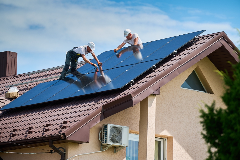 Workers building photovoltaic solar panel system on rooftop of house. Men technicians in helmets installing solar module with help of hex key outdoors. Renewable energy generation concept.
