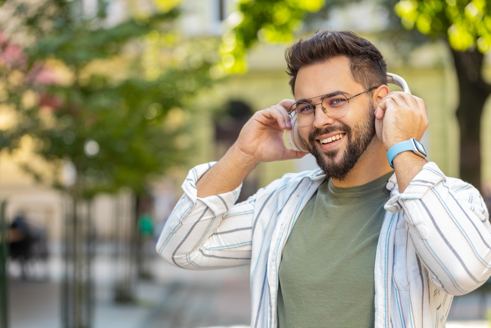 Joyful relaxed overjoyed young bearded man listening favorite energetic disco rock n roll music dancing outdoors. Adult guy taking off wireless headphones looking at camera smiling on city street.