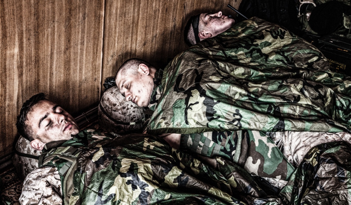 Tired U.S. marines resting at temporary base or camp, lying on floor in uniform and tactical ammunition, covered with poncho liners and sleeping bags, sleeping well after hard raid, exhausting mission
