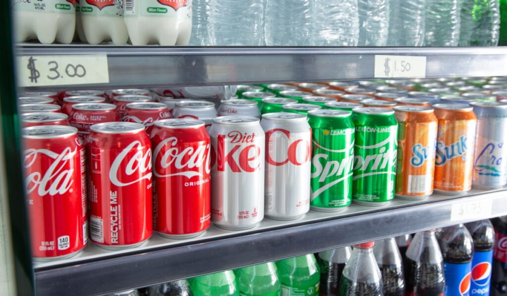 A view of a commercial refrigerator with a variety of popular soda brand cans, seen at a local convenient