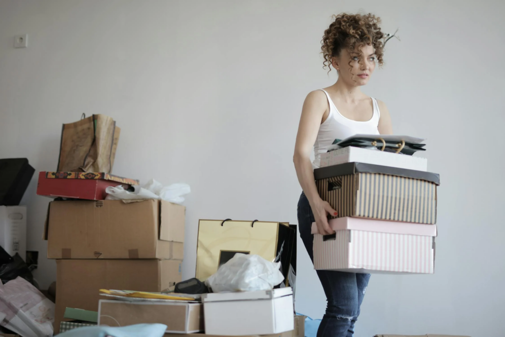A woman stands in a room filled with numerous boxes, surrounded by a cluttered yet organized environment.
