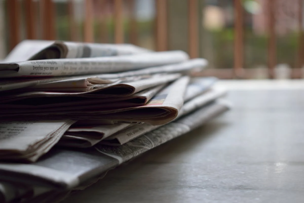 A stack of newspapers neatly arranged on a wooden table. Needing to be decluttered.