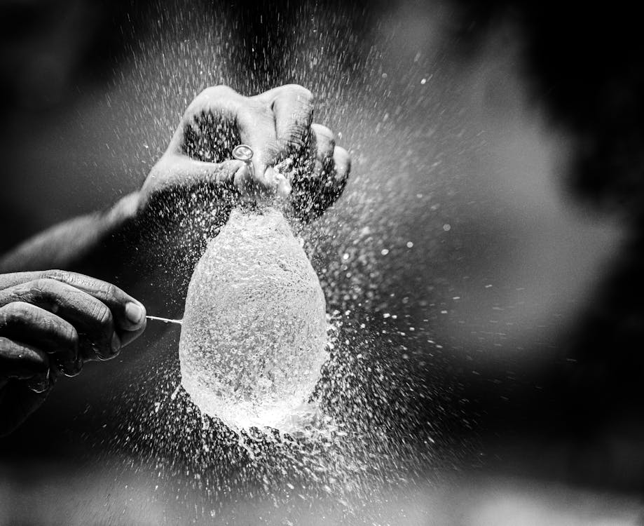 A close-up, black-and-white image of a hand popping a water balloon, with droplets bursting in every direction, reminiscent of the sudden release when popping a pimple.