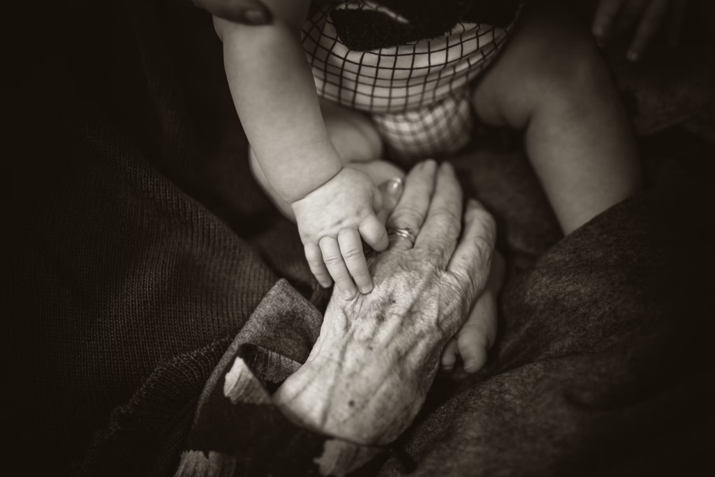 An elderly woman gently holds a baby's hand, symbolizing the connection between generations and the aging process.
