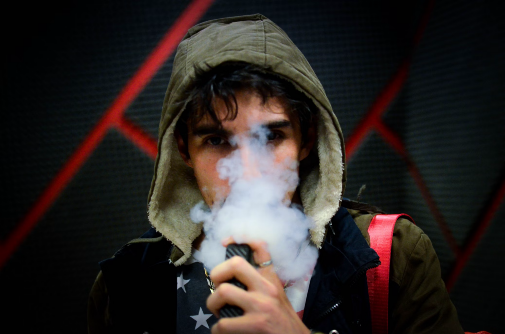 A young man in a hoodie smokes a vape, showcasing the concerning trend of vaping and its health risks.