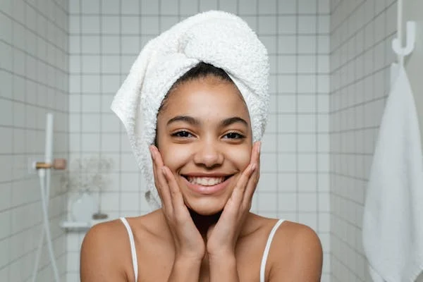 Girl in a bathroom, smiling with hands on her face and a towel on her head