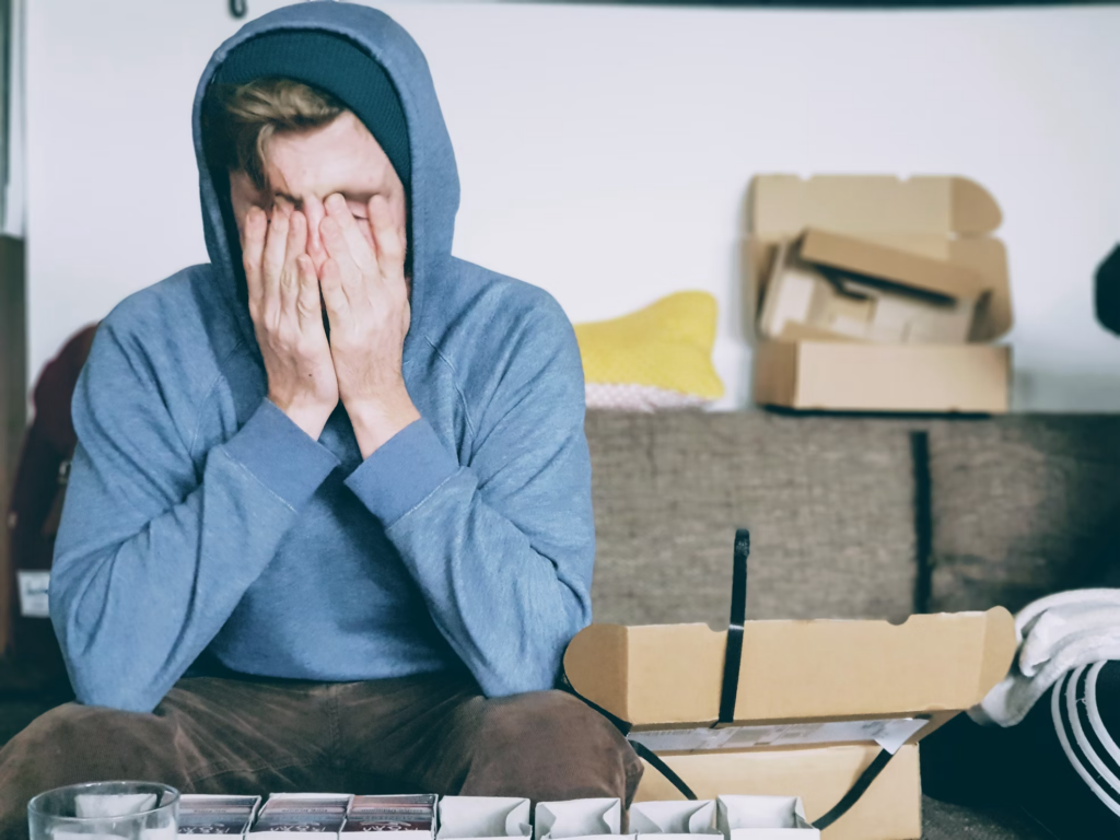 A man wearing a hoodie rests on a couch, with boxes around him, symbolizing the burden of depression-related fatigue.