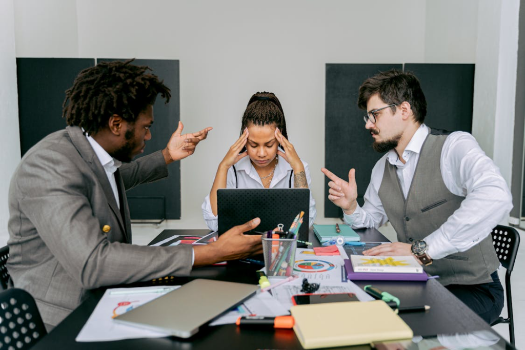 Three business individuals seated at a table. A woman in the background shows signs of irritation while using a laptop.
