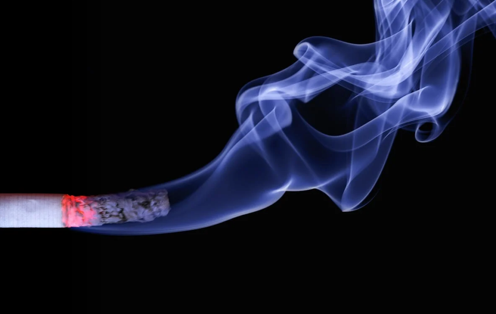 A lit cigarette emits a plume of smoke, creating a hazy atmosphere around it.