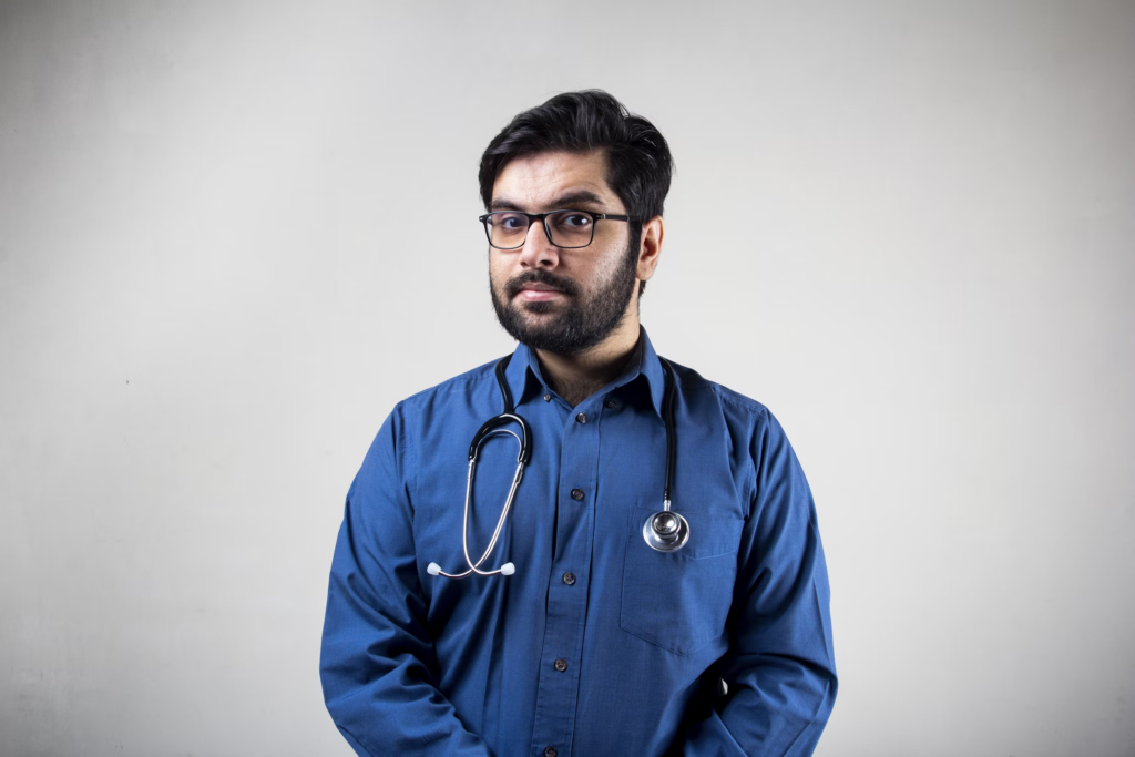 A male doctor wearing a blue shirt and glasses, standing confidently in a medical setting.
