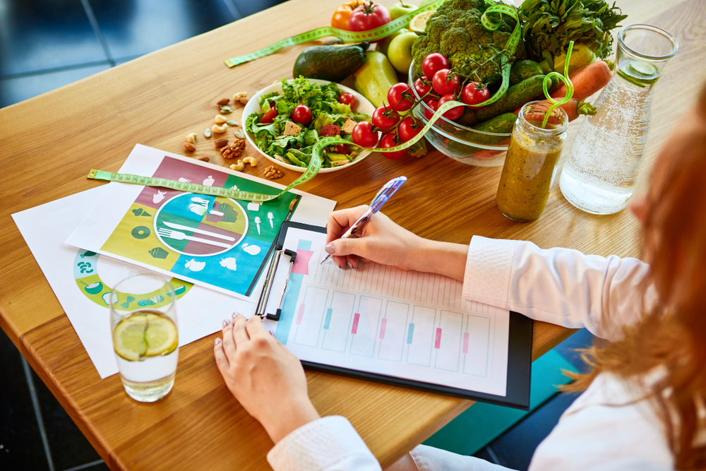 Woman dietitian in medical uniform with tape measure working on a diet plan sitting with different healthy food ingredients in the green office on background. Weight loss and right nutrition concept

