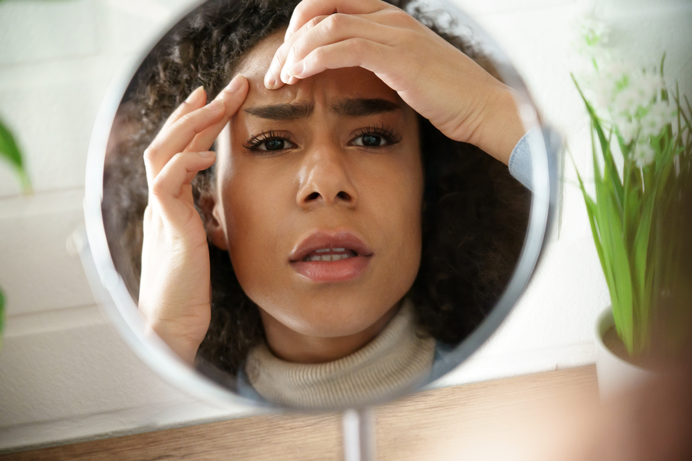 Worried african teen girl checking facial skin problem look in mirror. Young woman touch face squish pop zit pimple on forehead depressed by blackheads. Black skin care problem self care concept.

