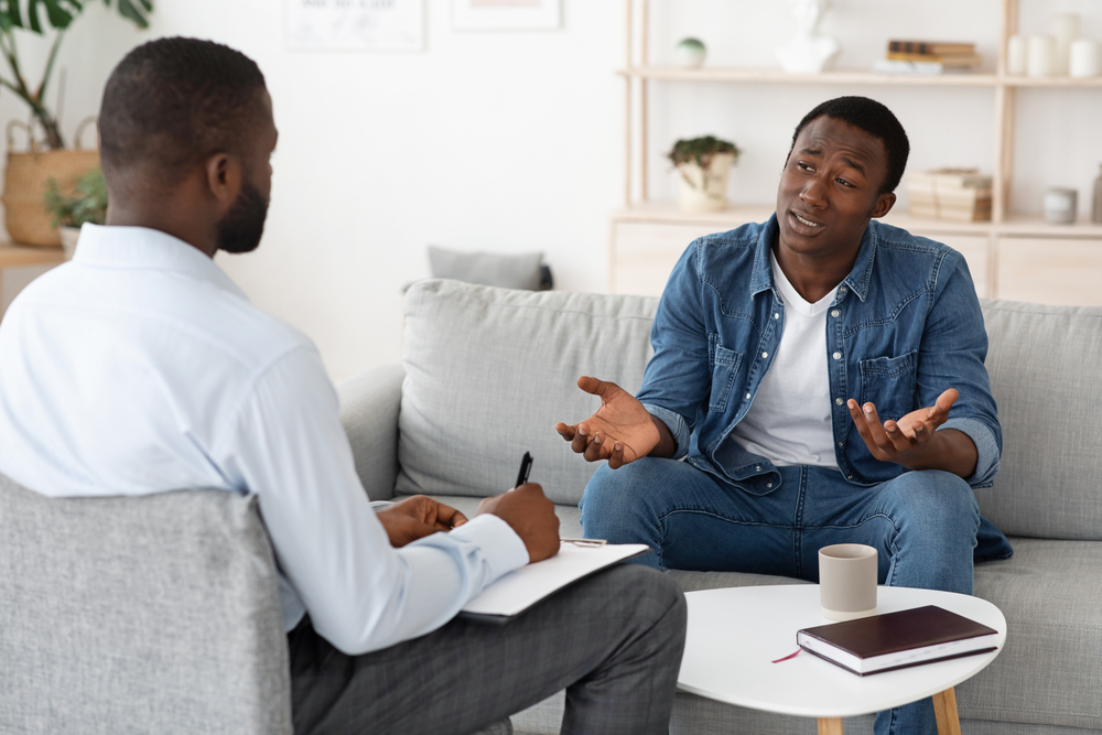 Stressed black man explaining his problems to psychologist at individual therapy session at office, copy space
