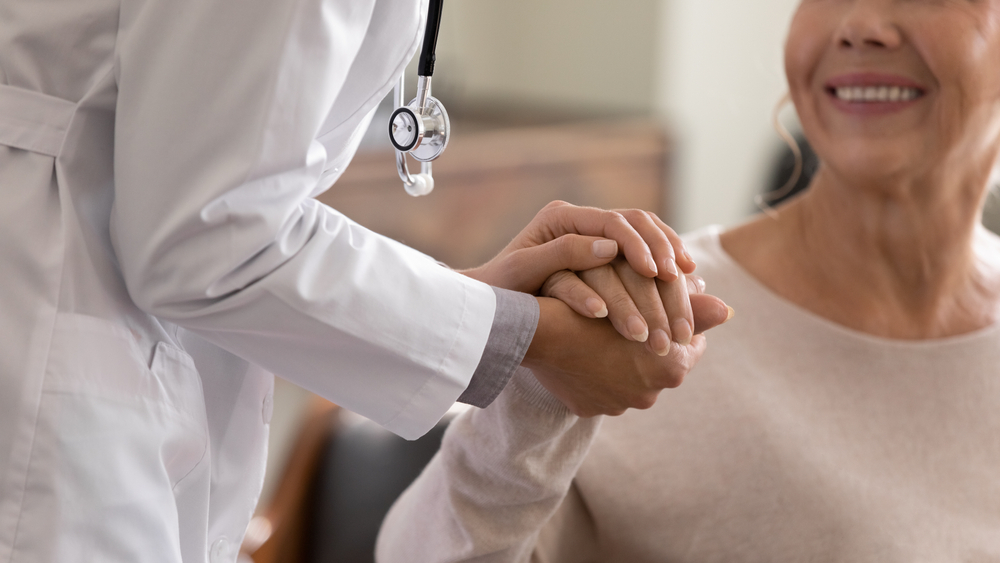 Doctor giving hope. Close up shot of young female physician leaning forward to smiling elderly lady patient holding her hand in palms. Woman caretaker in white coat supporting encouraging old person