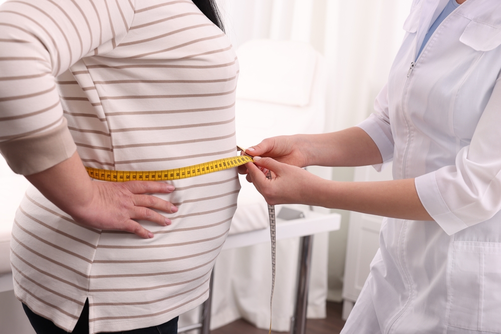 Nutritionist measuring overweight woman's waist with tape in clinic, closeup
