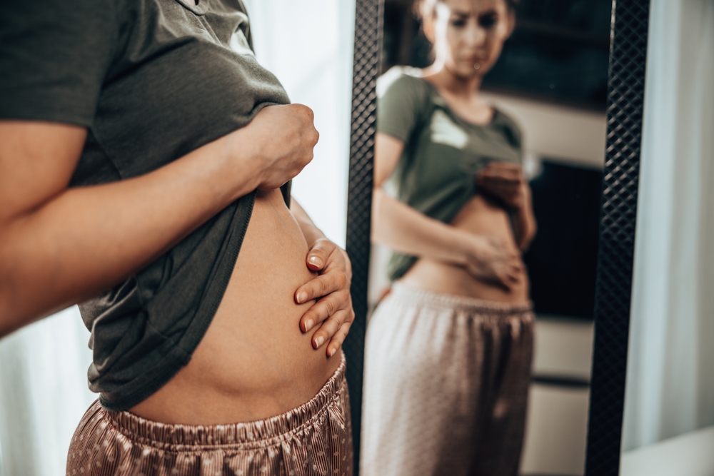 Unhappy young woman standing in front of a mirror and holding hands on her bloating stomach.
