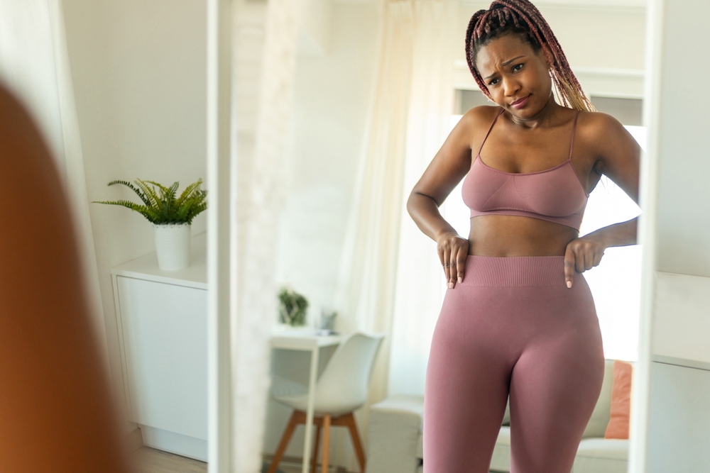 Sad and disappointed black lady standing in front of mirror and looking at reflection, unhappy about her body parameters. Slimming fail, unsuccessful weight loss
