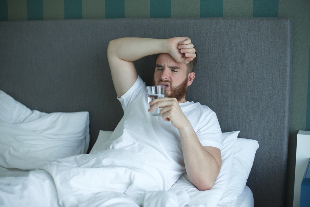 Young unhappy sick ill man in the bed in bedroom at home in the morning lying under white blanket and drinking pure fresh water from glass, suffering from headache, hangover