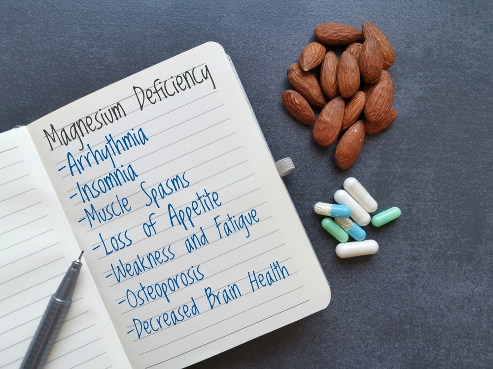 Symptoms and signs of magnesium deficiency written on the notebook, with almond and pills in background. Low magnesium level may cause weakness, fatigue, osteoporosis, muscle spasms, arrhythmia, etc.
